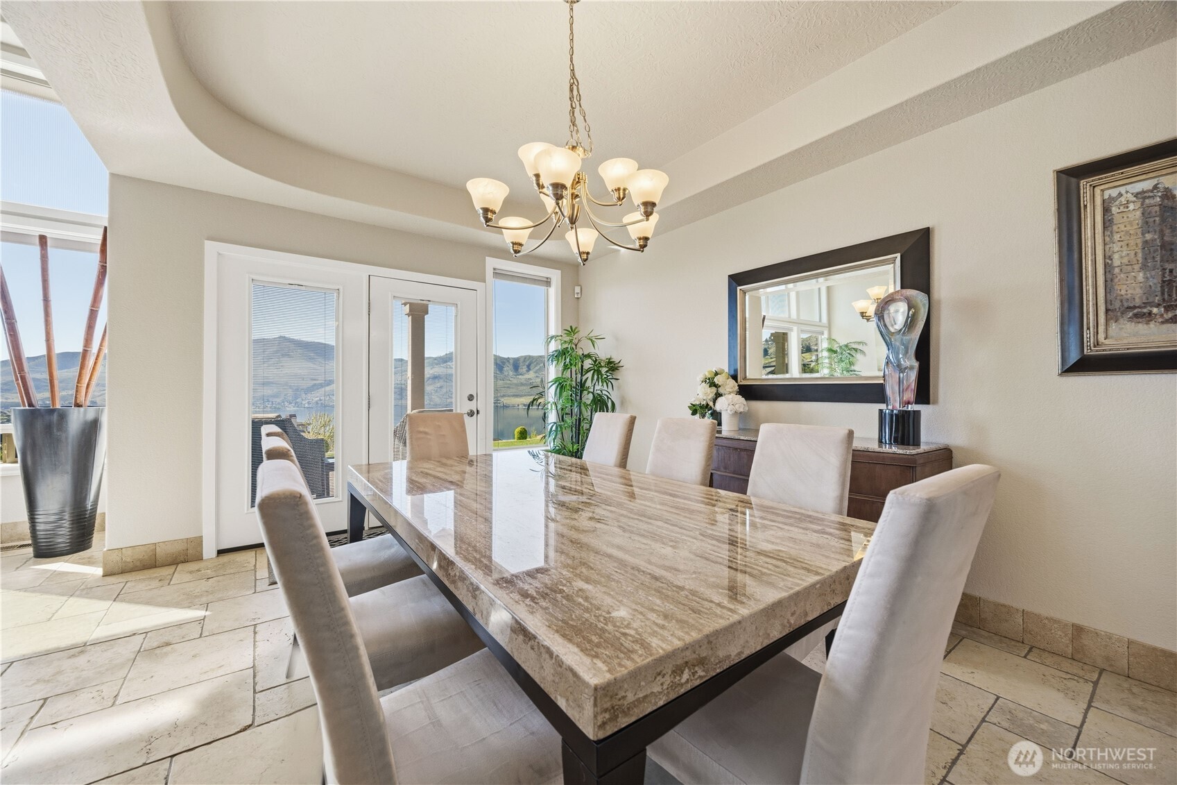 Elegant formal dining room with refined architectural details, including a tray ceiling and statement chandelier. Large windows and glass doors invite natural light while offering seamless access to outdoor living. Perfectly designed for hosting, this space blends sophistication and comfort for memorable gatherings.