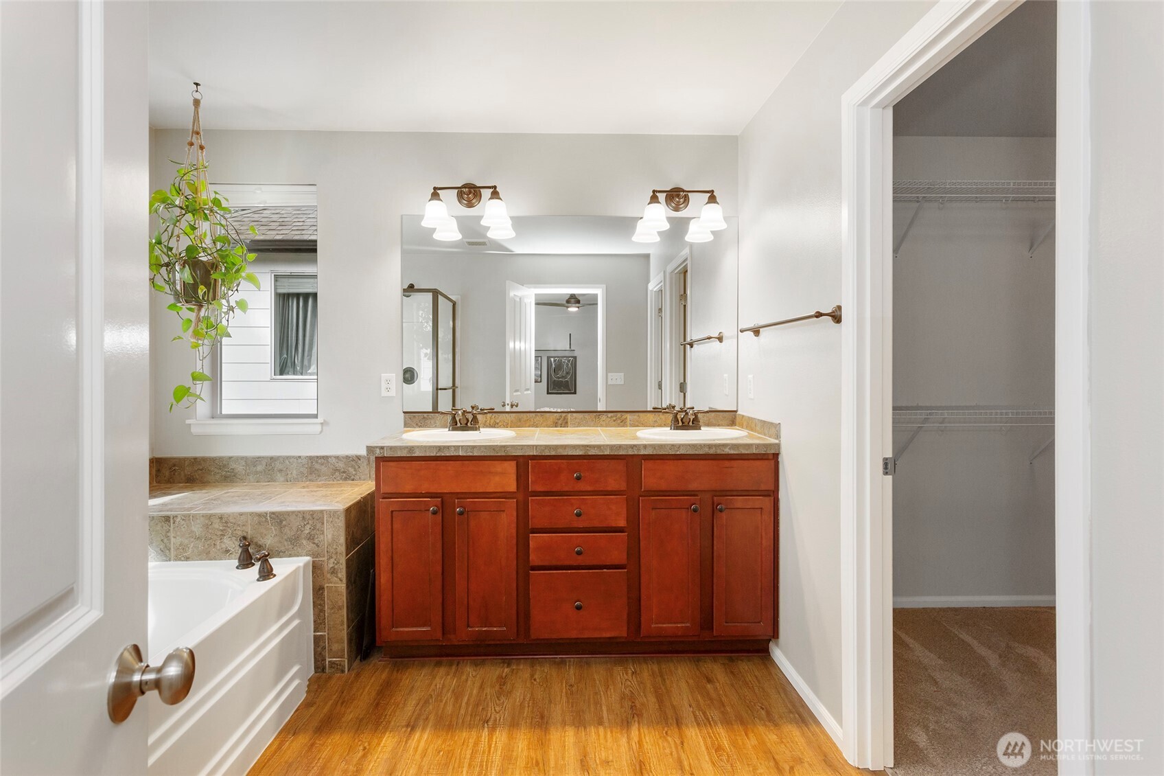 Primary bathroom with walk-in closet access featuring a spacious dual-sink vanity with warm wood cabinetry, generous counter space, and ample storage. Bright lighting, large mirror, and natural light create an open, airy feel. The door to the right leads to a massive walk-in closet, offering exceptional storage and a seamless, private flow within the primary suite.