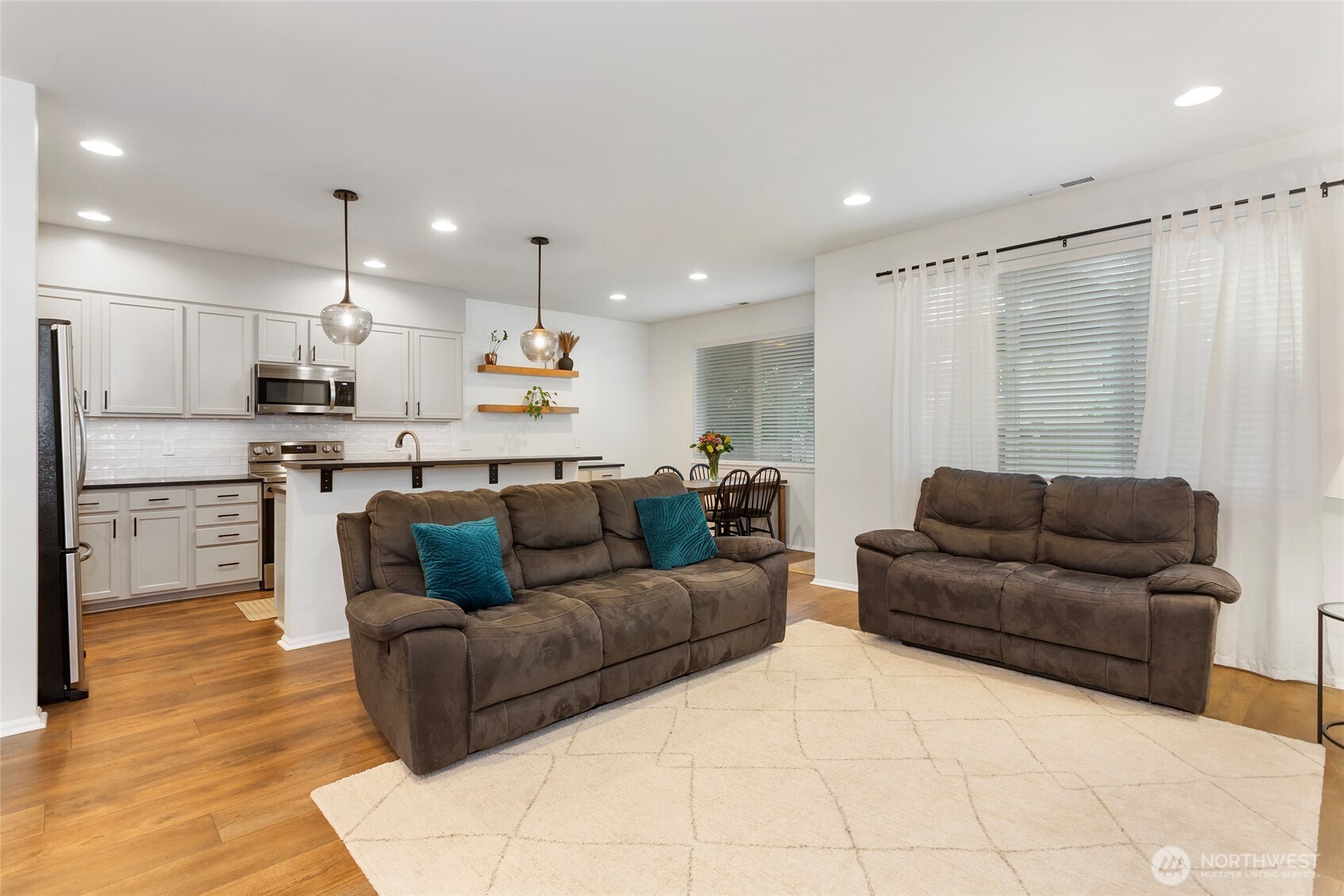 Bright open-concept living space featuring warm LVP floors, recessed lighting, and inviting natural light from oversized windows. The living room flows seamlessly into the upgraded kitchen, highlighted by a breakfast bar, modern pendant lights, and clean white cabinetry—ideal for both everyday living and entertaining.