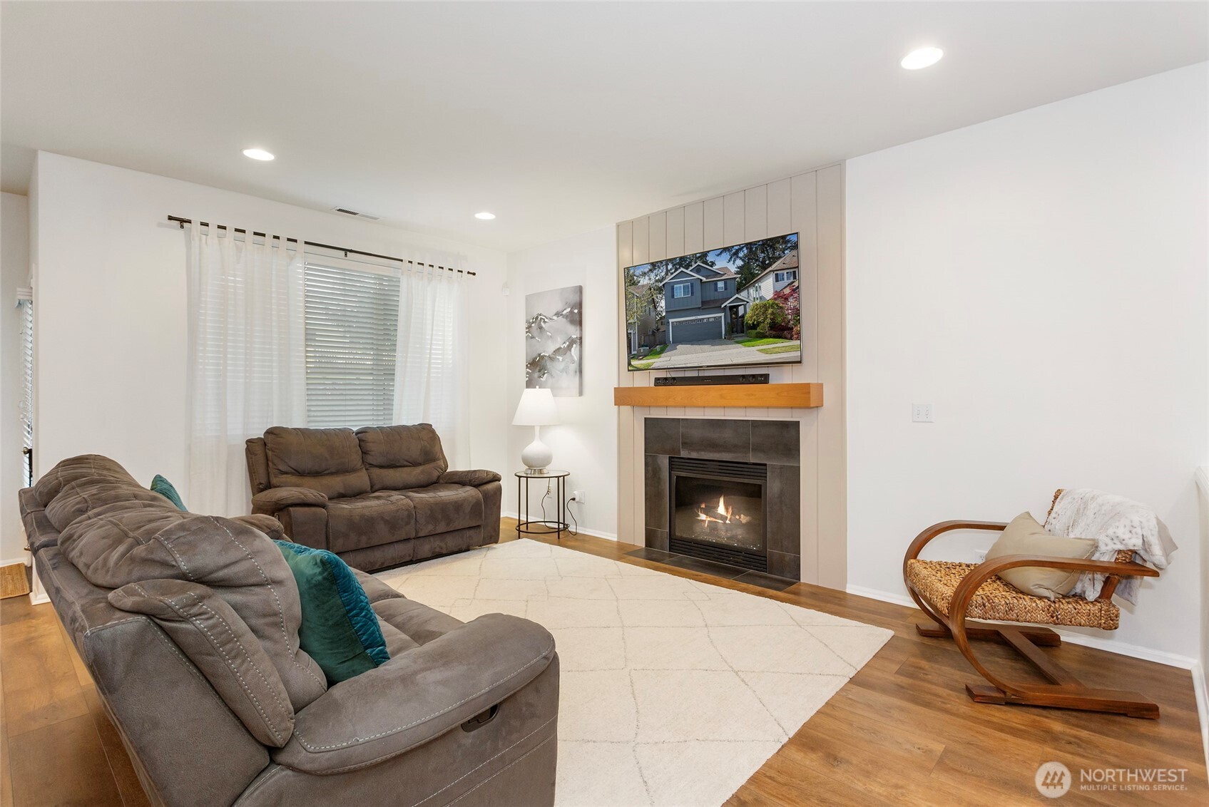 Contemporary living room centered around a sleek fireplace with modern tile surround and mantel, complemented by recessed lighting and warm LVP floors. Large windows bring in soft natural light, creating an inviting space perfect for relaxing or gathering.