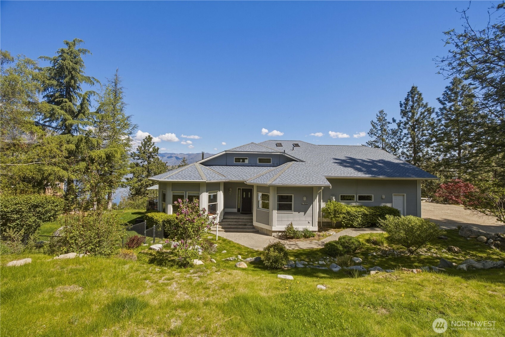 Surrounded by mature trees and lush green landscaping. A well-maintained lawn and natural vegetation frame the house, creating a private, peaceful setting.