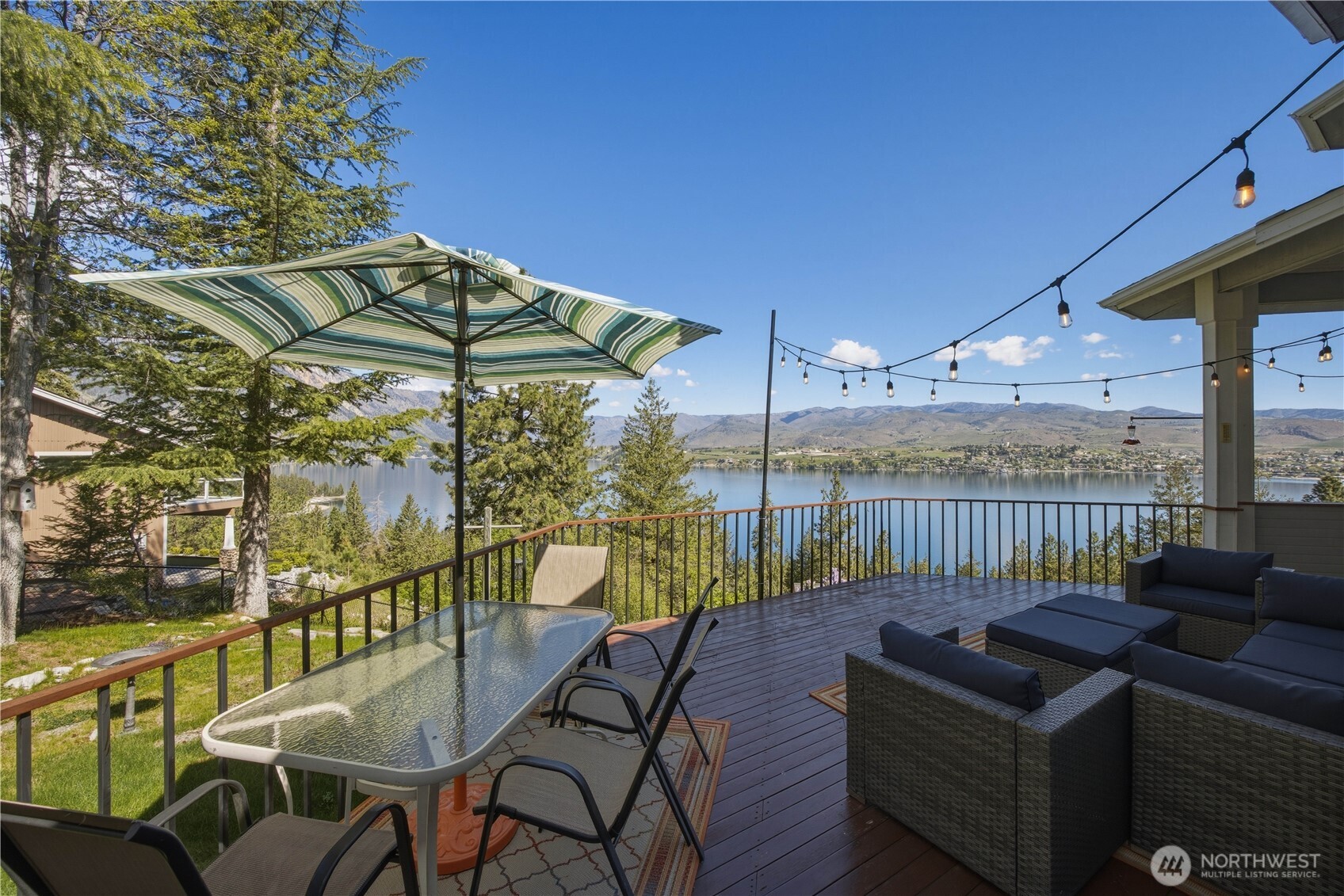 Outdoor deck with dining area, featuring a table and chairs, adjacent seating space, and railing that frames expansive lake and mountain views.