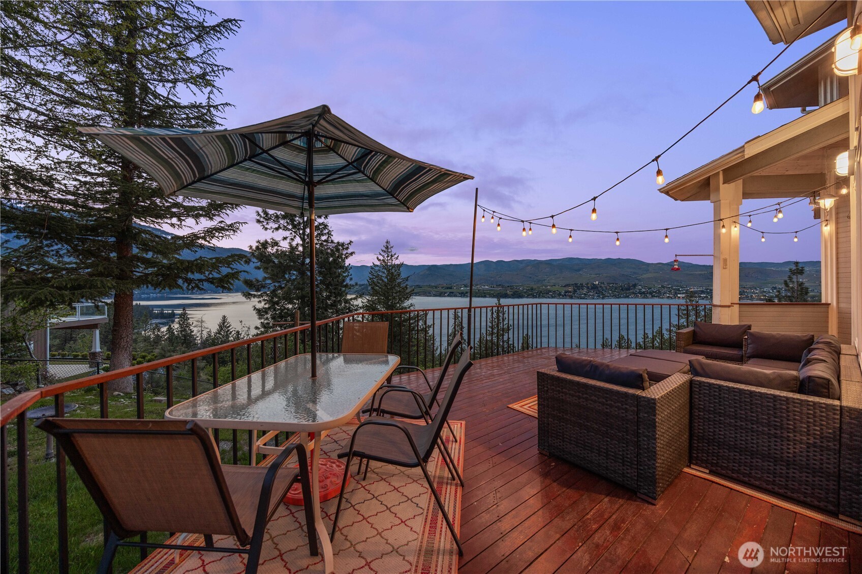 Spacious outdoor deck with seating area and dining space, overlooking expansive lake and mountain views. Surrounded by railings and mature trees, the setting offers a private, elevated vantage point for relaxing or entertaining.