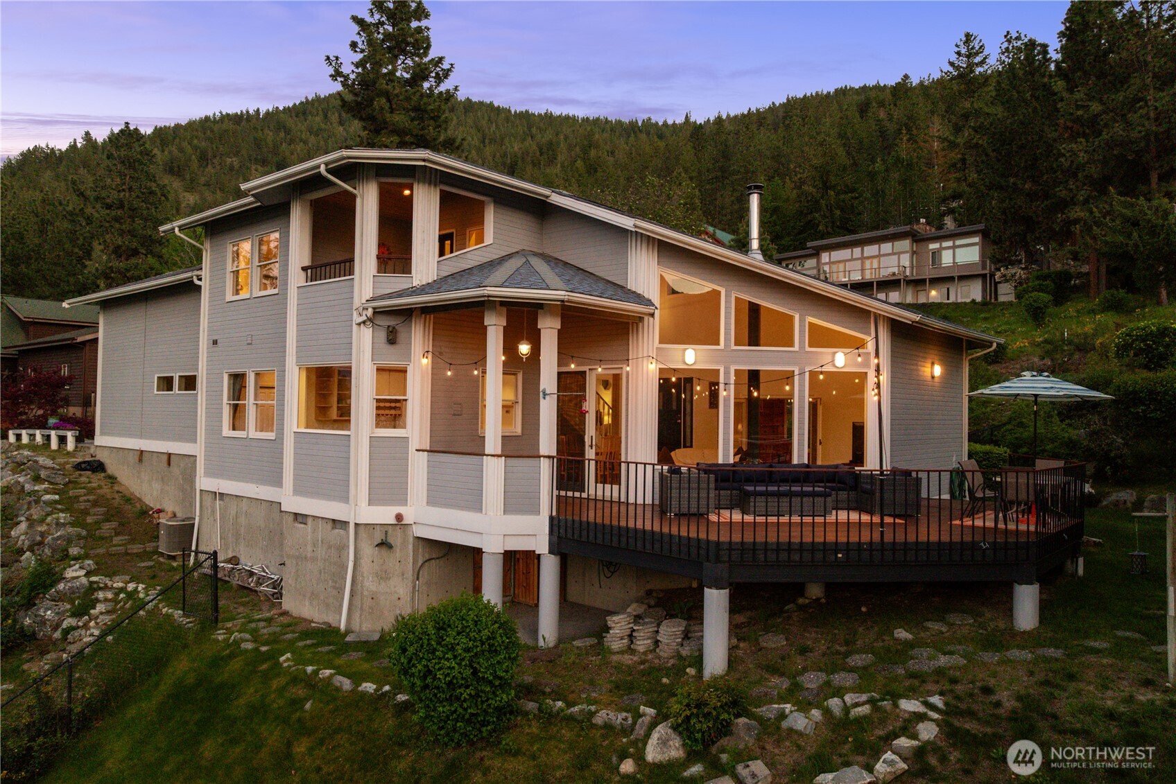 Elevated home with large windows, vaulted rooflines, and expansive wraparound deck. Surrounded by trees and natural landscape, the exterior showcases a warm, inviting design with scenic hillside views.