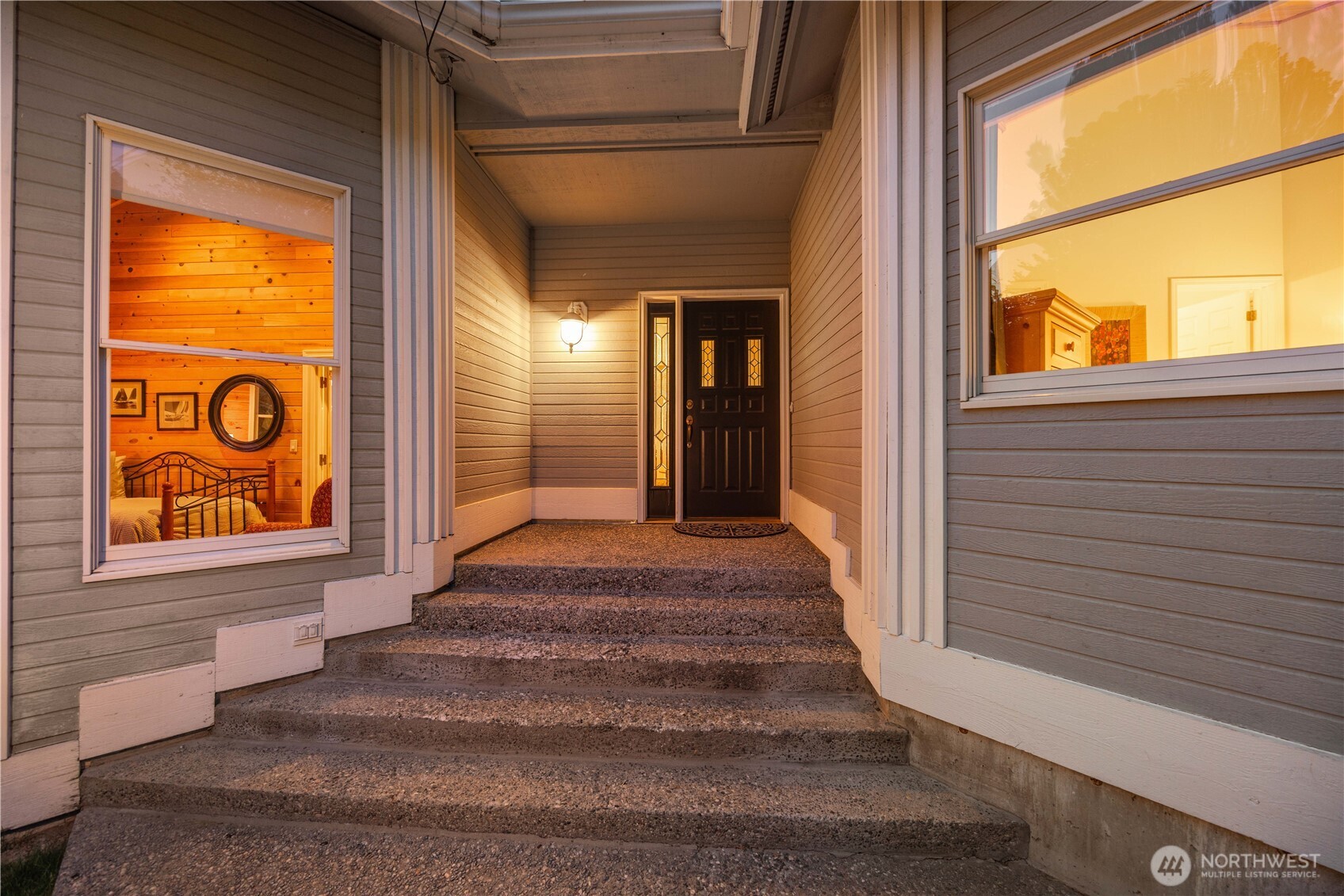 Covered entryway with wide steps leading to a recessed front door, framed by clean architectural lines and large windows. Warm interior lighting creates an inviting glow, highlighting the modern design and sheltered approach.