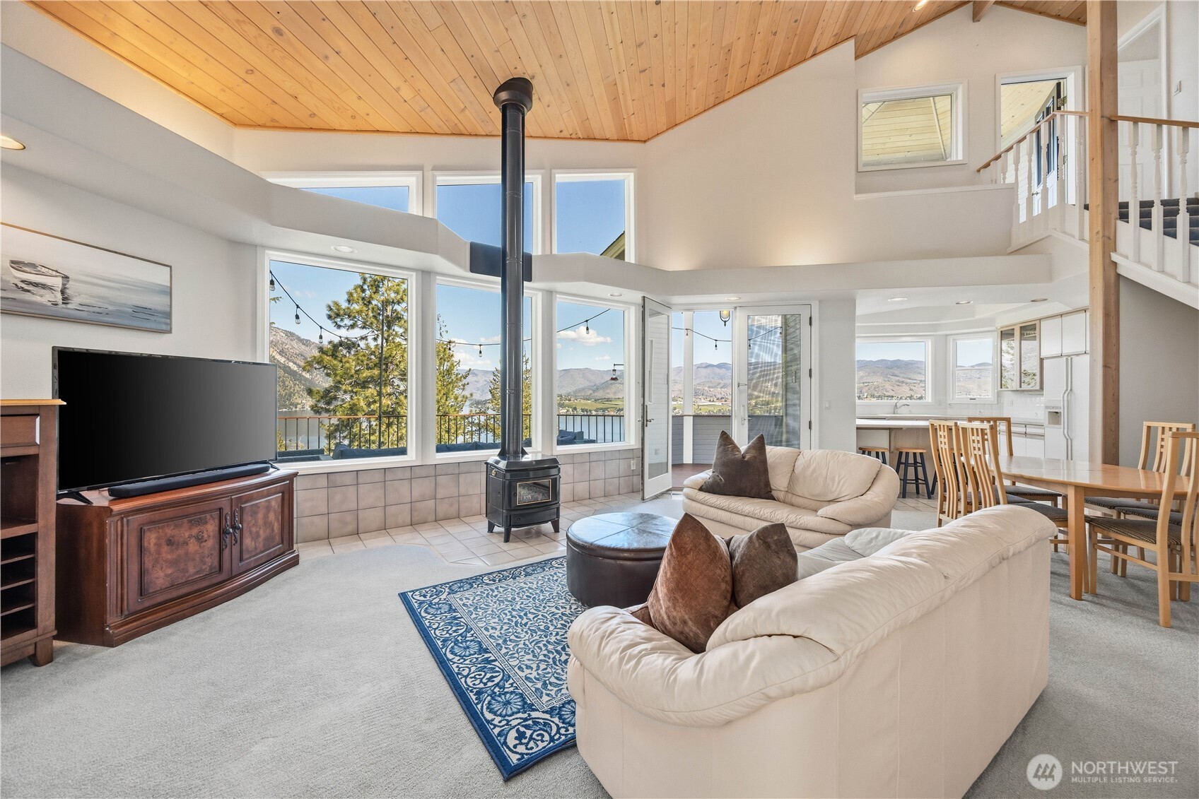 Open living area with vaulted ceiling, expansive windows, and a central freestanding stove. The space features a comfortable seating area, and an airy layout that flows into the adjacent kitchen.