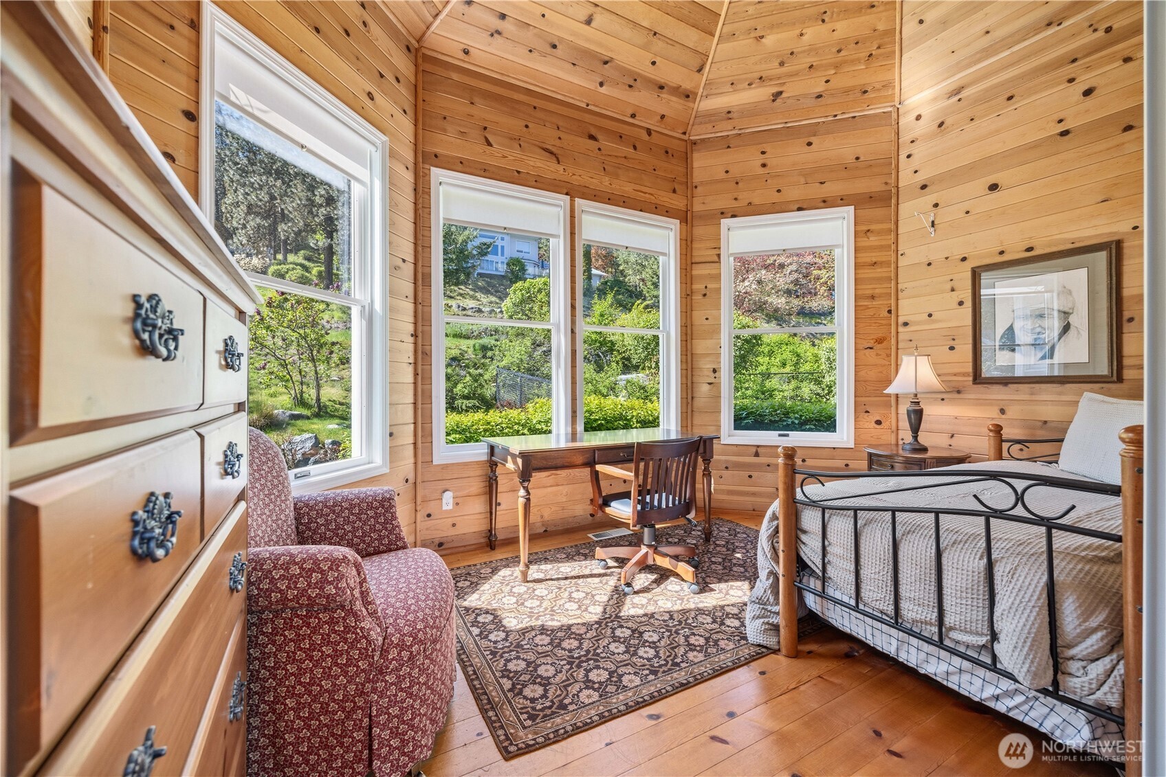 Cozy sitting area with warm wood walls and ceiling, surrounded by large windows that bring in natural light and scenic views. The space features comfortable seating, and is ideal for office or additional sleeping space if needed.