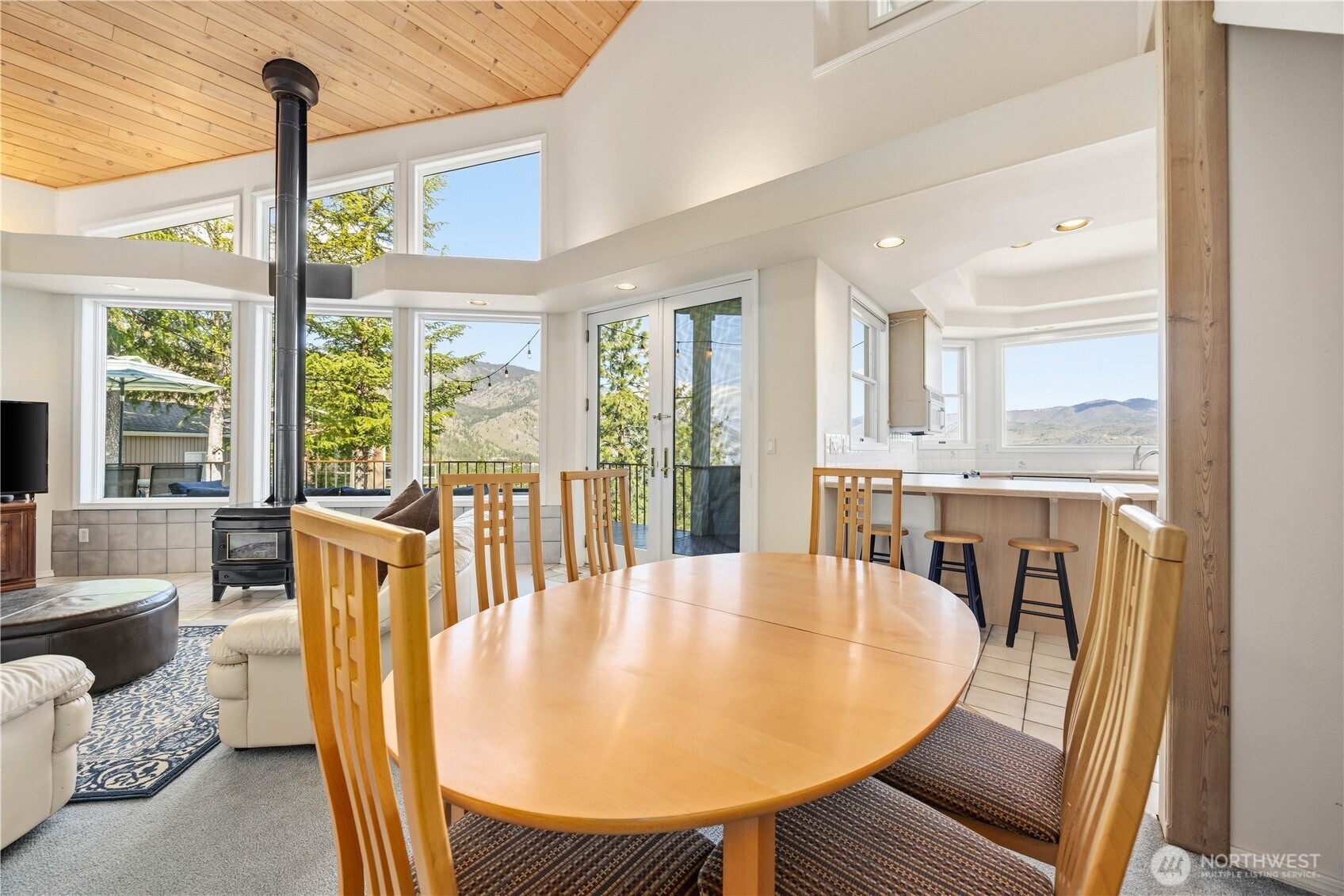 Open dining area with a round wood table and chairs, positioned between the living room and kitchen. Large windows and sliding glass doors fill the space with natural light, highlighting the vaulted wood-paneled ceiling and seamless flow between living areas.