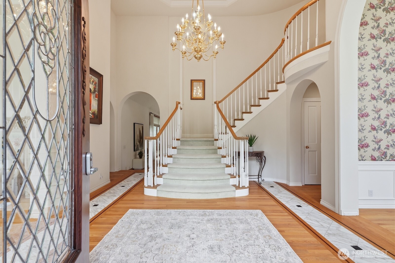 The striking castle stone entry, adorned with a rounded leaded glass door, opens to a breathtaking grand foyer featuring soaring ceilings and elegant wood-wrapped archways that seamlessly connect the formal living and dining spaces.