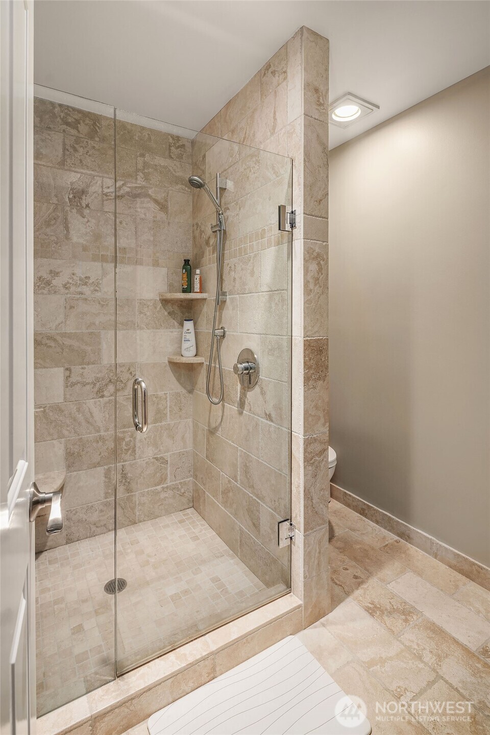 The en-suite bath delivers a spa-like experience in warm travertine — floor-to-ceiling stone tile wraps a generously sized walk-in shower enclosed in frameless glass, with a handheld shower, built-in corner shelving, and coordinating tile floors throughout. A quiet luxury that guests will not soon forget.