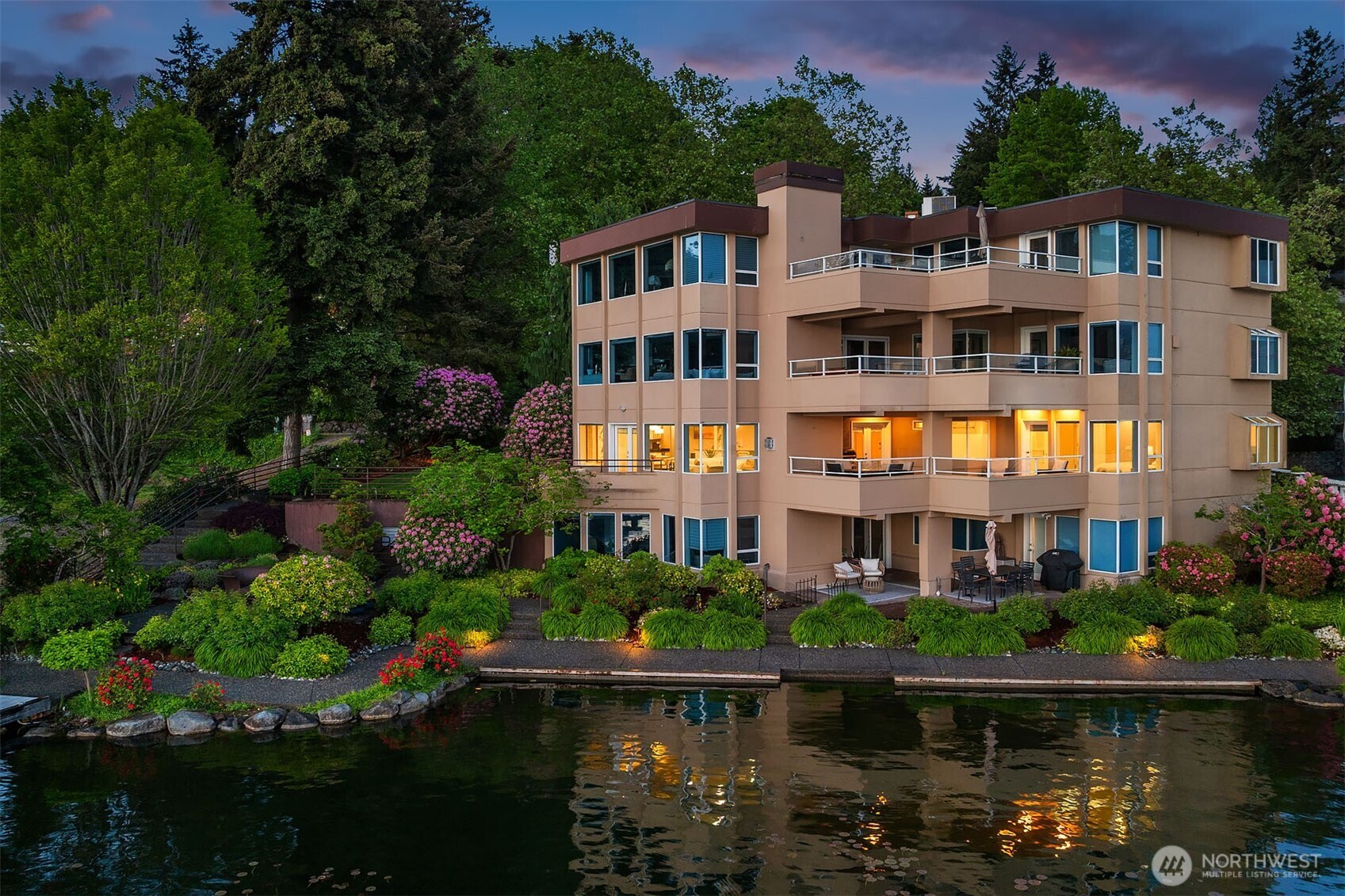 An entire floor of a four-unit waterfront building, glowing against the twilight sky with walls of glass, multiple decks, and spectacular blooming landscaping cascading to the water's edge. Level entry from the street, a lush lawn at the side, and just steps to the gated dock below. The privacy and presence of a single-family home, without compromise.
