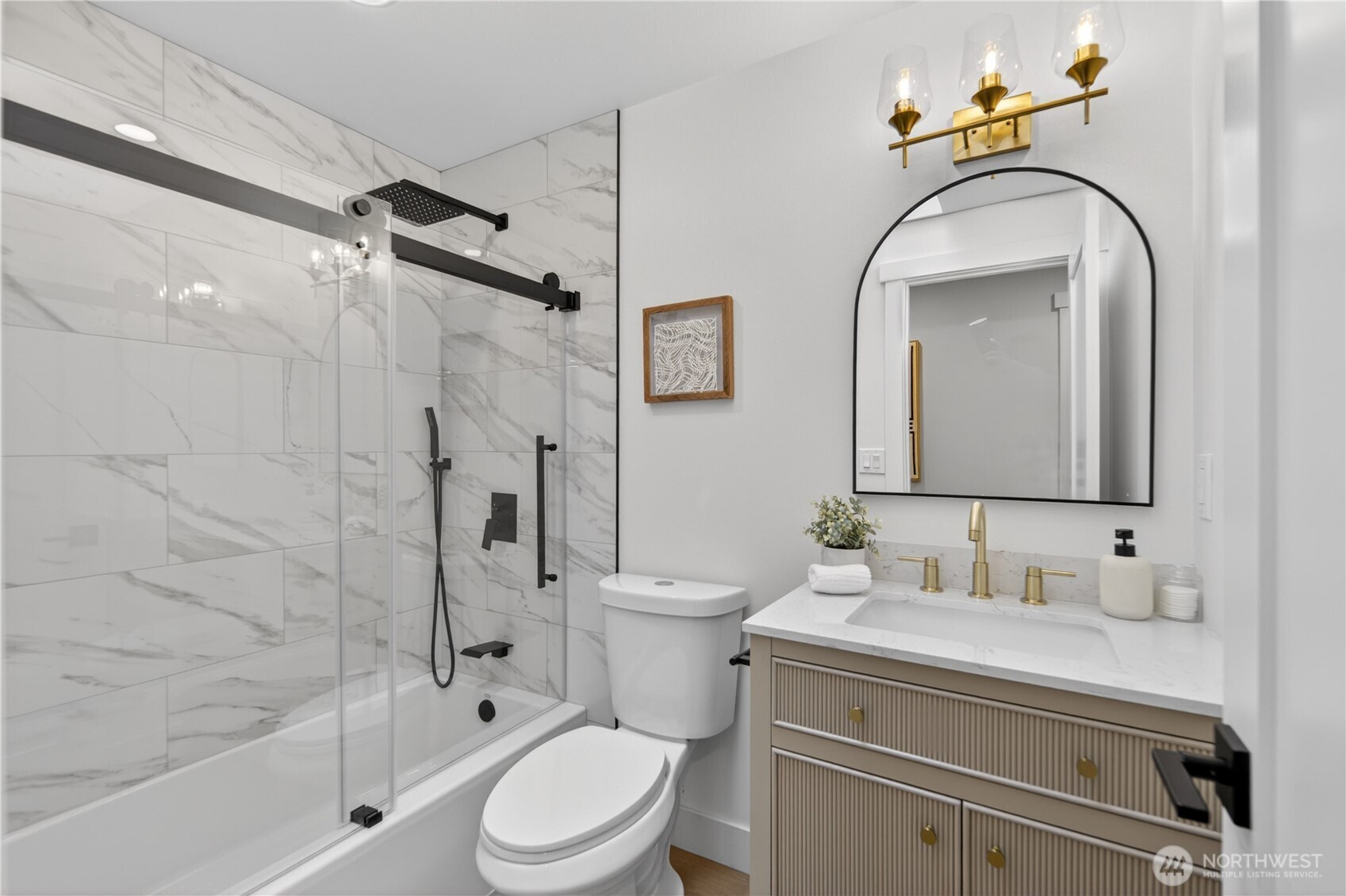 Bright white walls and a skylight bring natural light into this bath, creating a space that feels bright, elevated, and beautifully refreshed. Glass shower doors showcase marbled shower tile that echoes the kitchen finishes, giving the home a cohesive designer feel, while black shower hardware, a rainfall shower head, and handheld sprayer add modern comfort. A fluted furniture-style vanity brings texture and generous storage, topped with white quartz that reflects the kitchen’s clean, refined surfaces. Gold-tone swan-neck faucets, an arched black-framed mirror, and elegant glass-shade vanity lighting complete the room with warmth and polish.