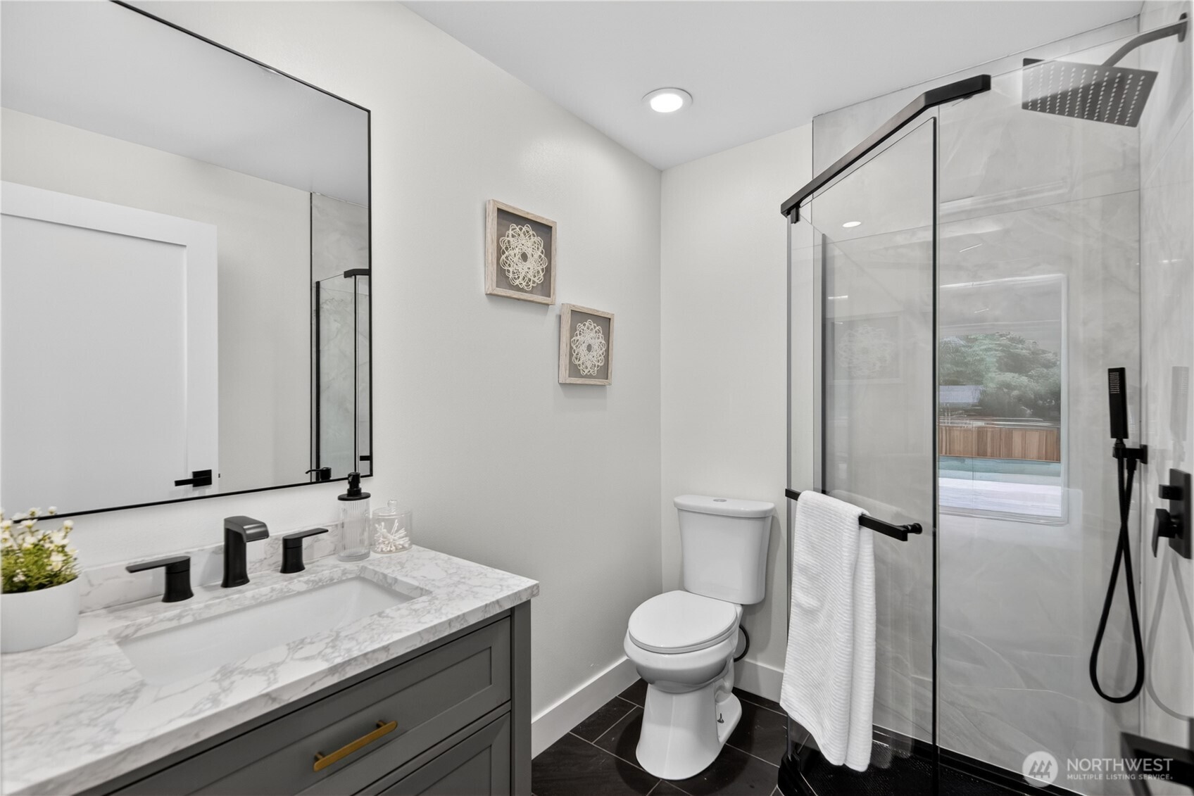 An all-new three-quarter bath completes the lower level with a beautifully refreshed finish. The glass-walled walk-in shower is wrapped in floor-to-ceiling marbled tile, with black-tone fixtures, a rainfall shower head, and handheld sprayer for modern comfort. The vanity adds both beauty and function, with a generous cabinet, marbled quartz counter, and waterfall-style faucet that echo the shower’s refined finishes. A large mirror expands the room visually, while warm recessed lighting and a window for natural light keep the space bright and welcoming. The tiled floor adds a clean, finished touch for guests or extended family.