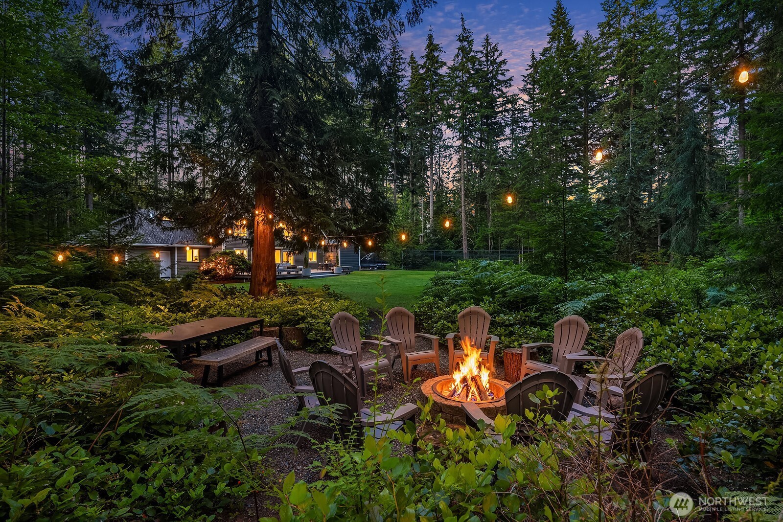 A charming stone fire pit gathering area anchors the backyard, offering a serene outdoor retreat with ample flat, usable acreage and the warm glow of the home visible through the trees.