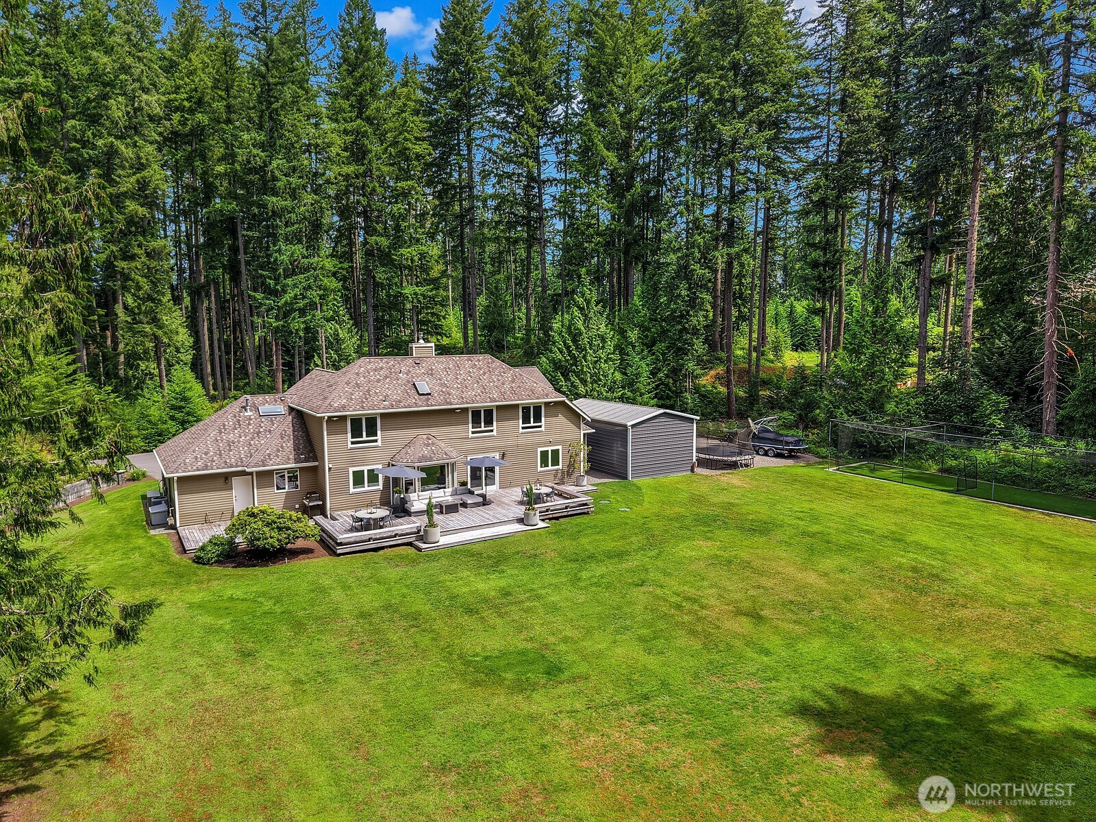 An aerial view showcases the expansive, flat usable backyard with lush green lawn, Garapa hardwood deck, and mature evergreen tree line providing exceptional privacy on all sides.