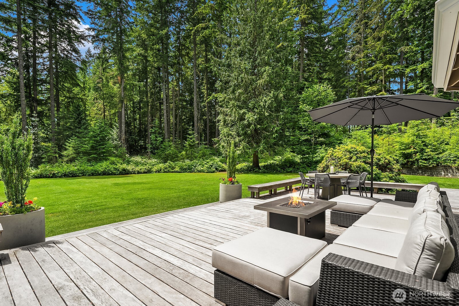 The Garapa hardwood deck offers a generous outdoor living space with room for lounging and dining, overlooking the lush, flat backyard framed by towering evergreens for total seclusion.