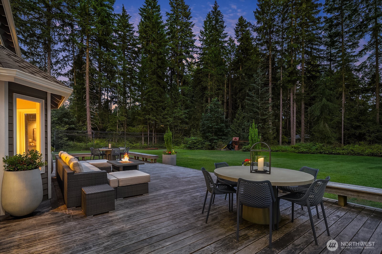 The deck and patio area come alive in the evening with warm ambient lighting, offering an inviting outdoor dining and lounge space perfectly framed by the towering evergreens of the private backyard.
