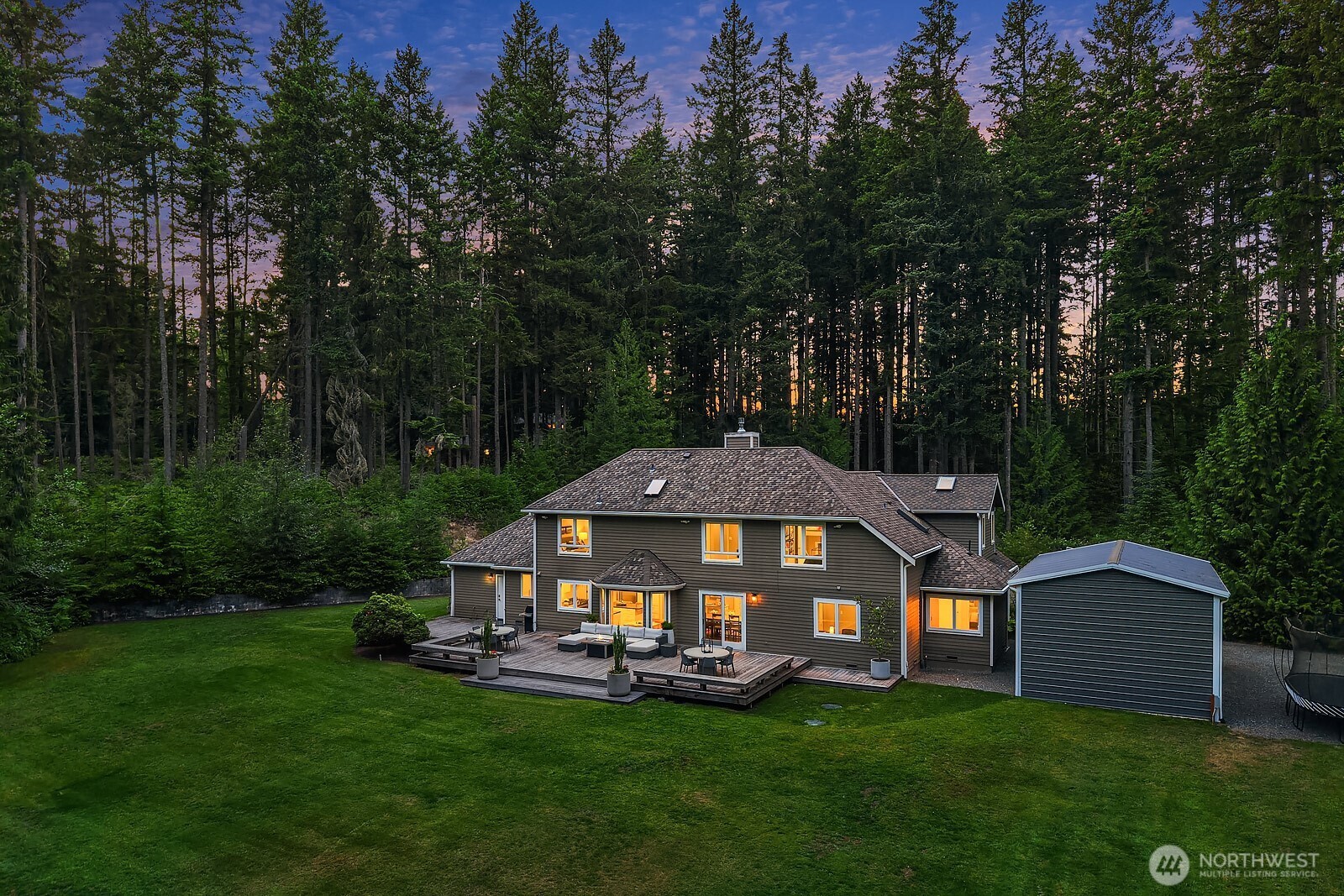 This aerial dusk view captures the full scope of the property, highlighting the beautifully lit home, expansive deck, detached shop, and sweeping flat lawn all set within a stunning evergreen backdrop.