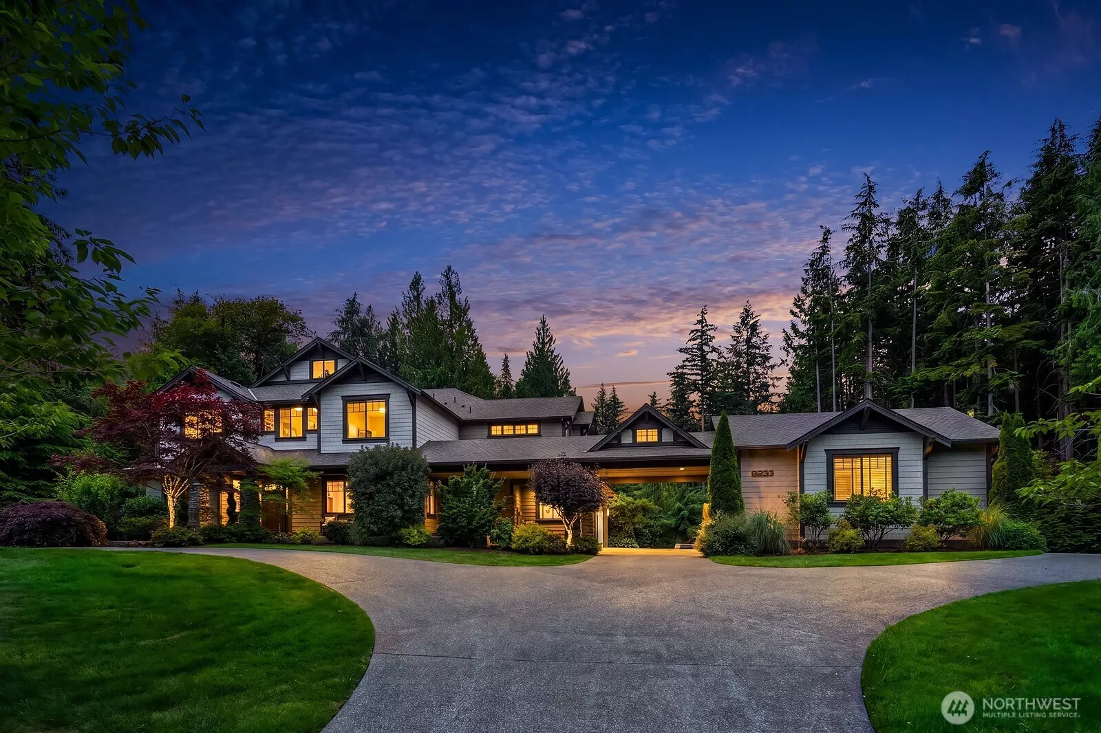 Stunning craftsman-style estate set against a lush Pacific Northwest backdrop, showcasing the home's sweeping wrap-around driveway and beautifully illuminated exterior at dusk. Nearly 2.5 private acres of mature trees and manicured landscaping frame this 4,710 sq ft showpiece in the coveted Meadowood community.