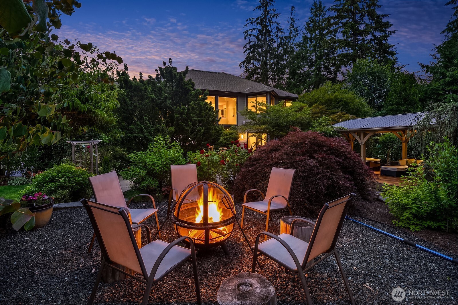 The raised gravel fire pit area offers a magical evening gathering spot, with warm flickering light set against the beautifully illuminated home and lush mature landscaping creating an unforgettable outdoor ambiance.