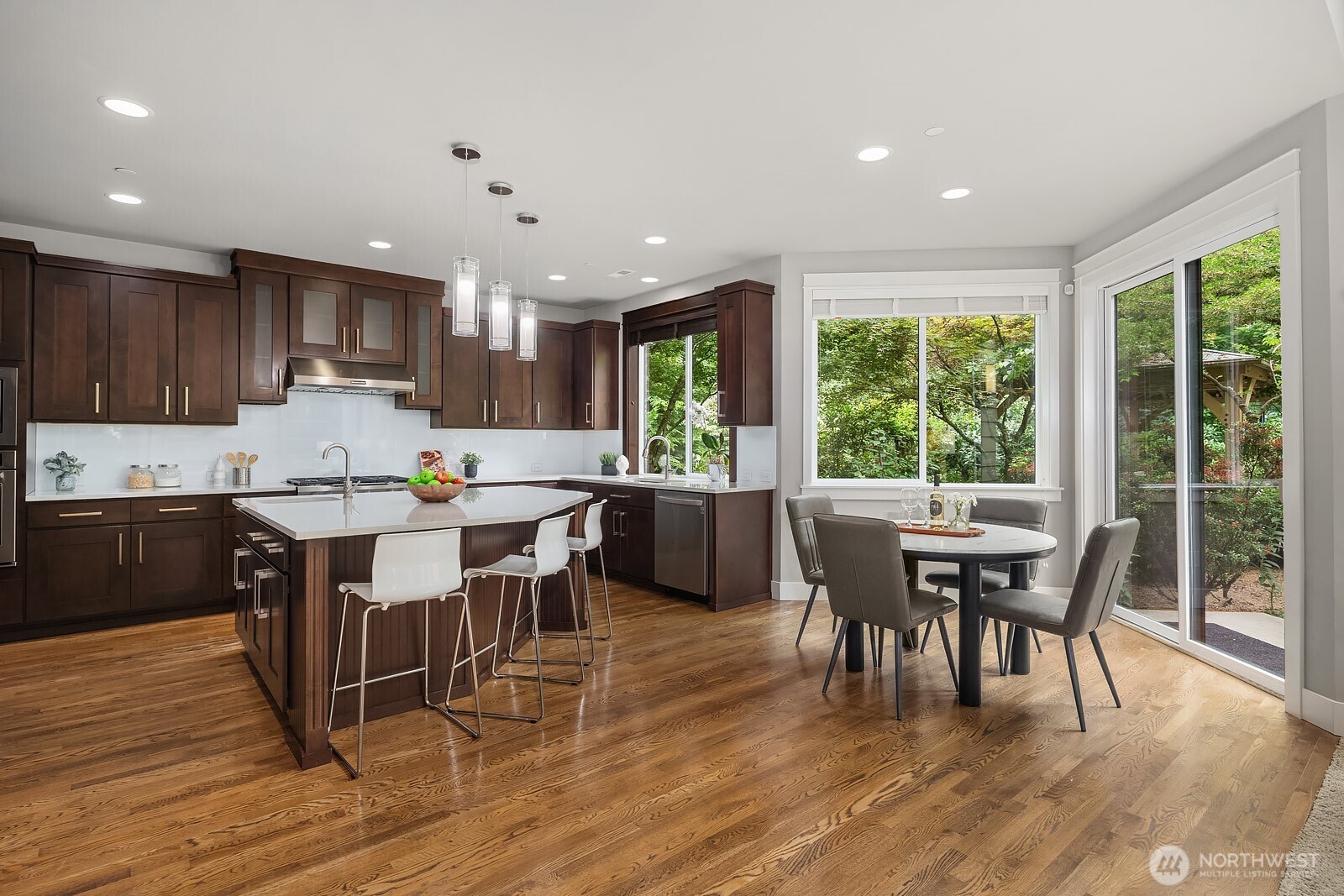The chef's kitchen impresses with rich dark cabinetry, premium appliances, and a generous island with seating, all anchored by warm hardwood floors. A sun-drenched dining area just beyond offers the perfect spot for casual meals with tranquil garden views.