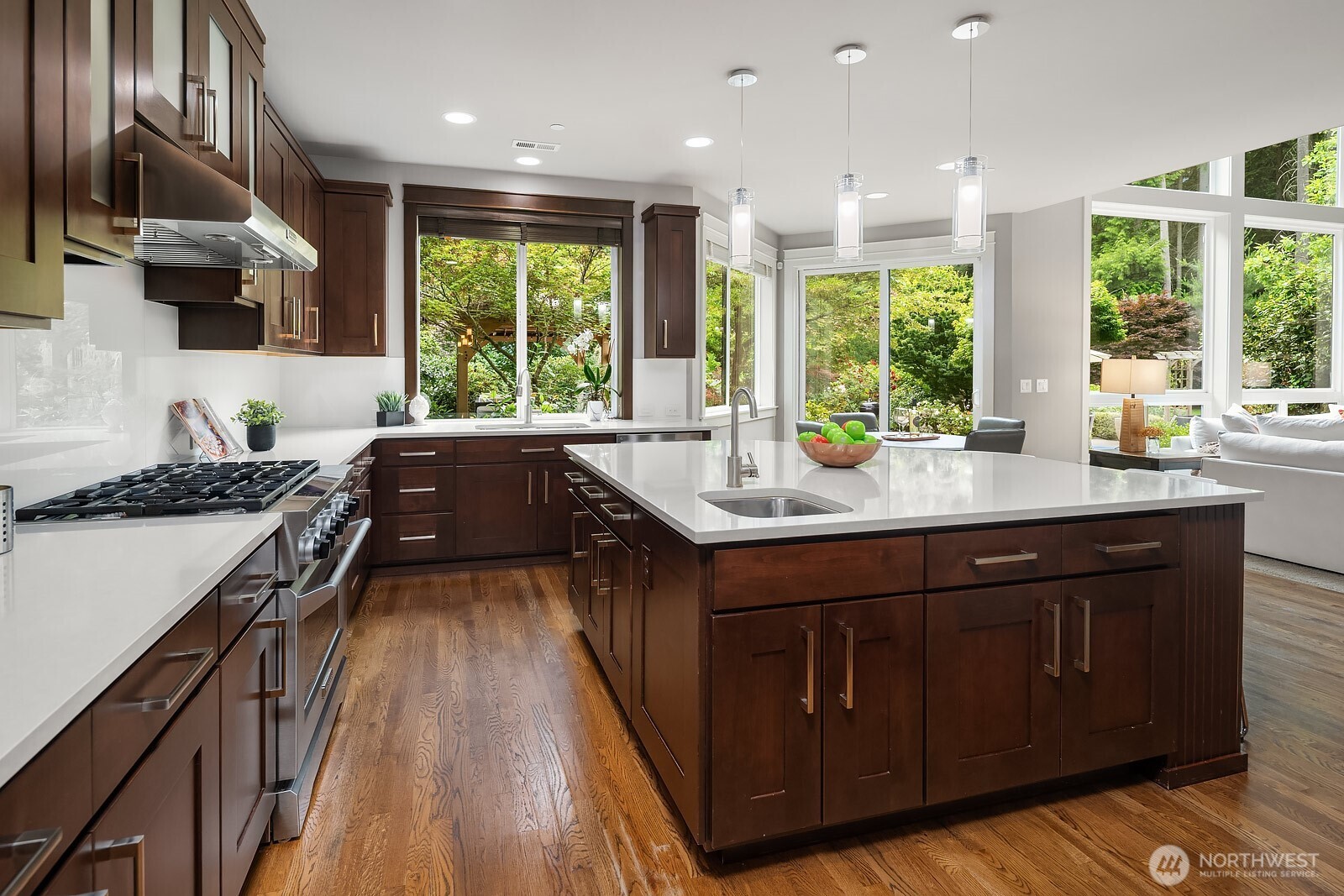 This expansive kitchen layout showcases dual islands, extensive dark cabinetry, and gleaming countertops that offer exceptional prep and storage space for the most discerning home chef. Wraparound windows bathe the space in natural light while framing the lush greenery of the surrounding grounds.
