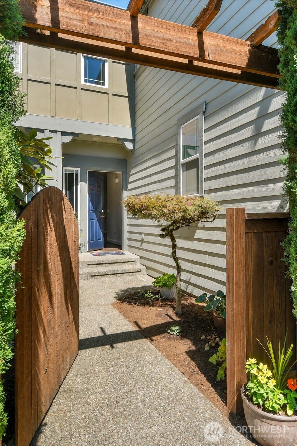 Aggregate walkway through gated entrance framed with wooden, decorative pergola, and courtyard with separate garden areas for each home. Paneled Quaker entry door with Eufy® Video doorbell, security deadbolt lock and sidelight window with shade.