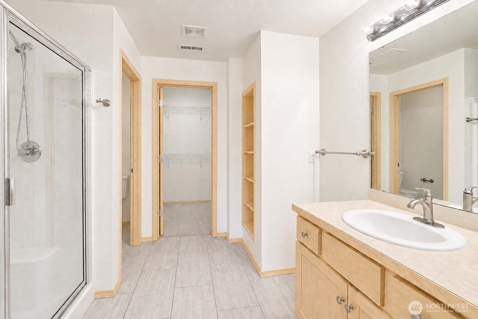 Primary ensuite bath. Tile floor. Vanity with Shaker-style cabinet, Formica®-style countertop, vanity mirror with Chrome backed globe vanity lights, newer sink and brushed nickel faucet. Brushed nickel towel bars. Faux-tile wraparound shower with Chrome-framed glass door. Alcove with linen shelving. Primary closet with wire shelving, neutral carpet. Watercloset room with tile floor, newer American Standard® watercloset.