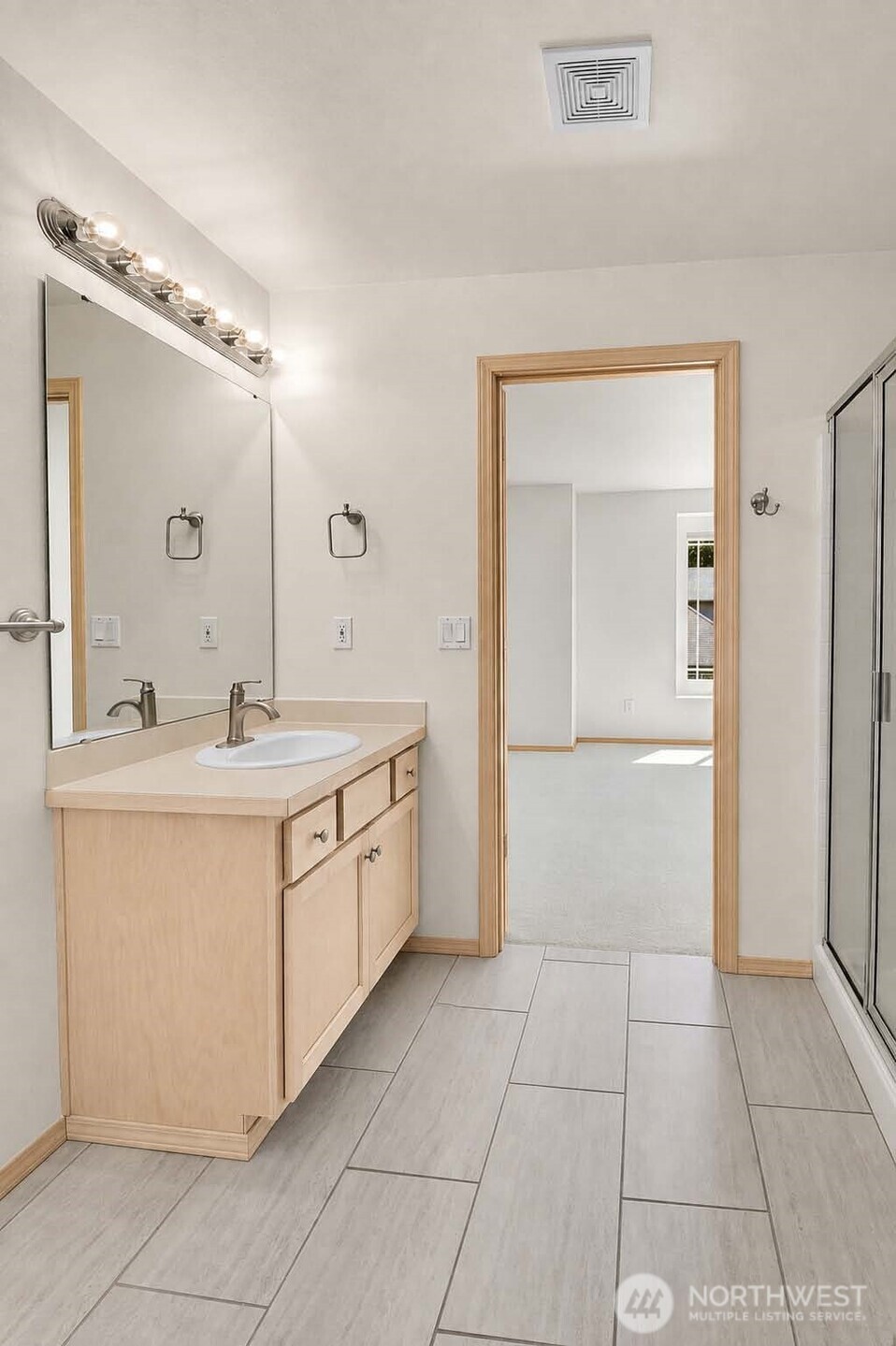 Primary ensuite bath. Tile floor. Vanity with Shaker-style cabinet, Formica®-style countertop, vanity mirror with Chrome backed globe vanity lights. Brushed nickel towel bars. Faux-tile wraparound shower with Chrome-framed glass door. Alcove with linen shelving. Primary closet with wire shelving, neutral carpet. Watercloset room with tile floor, American Standard® watercloset.