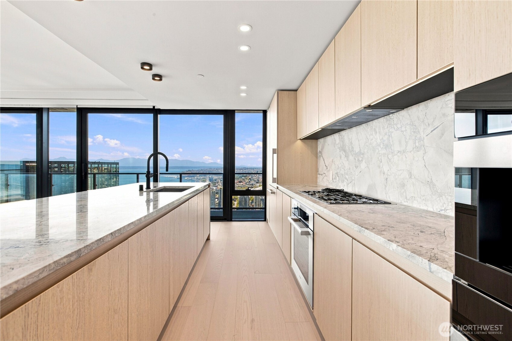 The definition of a luxury kitchen, anchored by a grand Italian marble island with sculpted waterfall edges and appointed with a complete suite of premium Miele appliances—from a generously sized wine refrigerator to an integrated espresso and drip coffee system. Natural wood B&B Italia cabinetry echoes the warm tones of the hardwood flooring, creating a seamless composition of refined materials, craftsmanship, and effortless functionality.