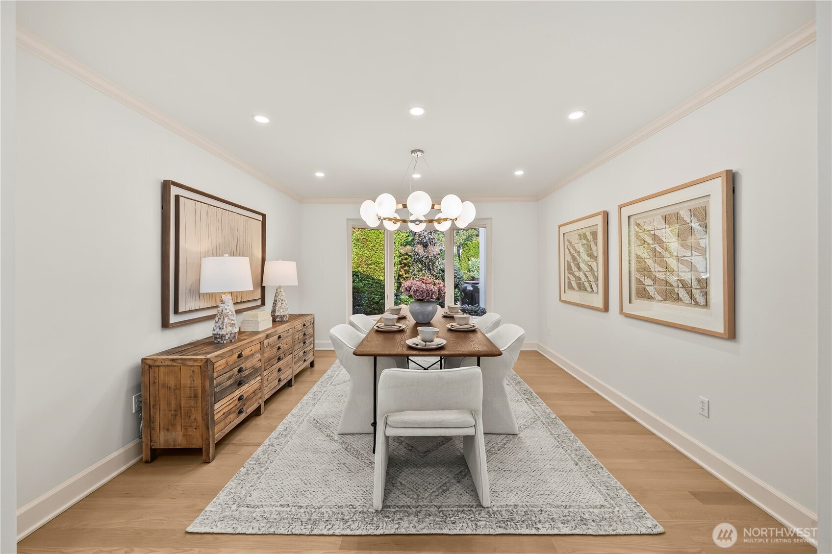 A sophisticated dining room framed by expansive windows and lush garden views. A contemporary statement chandelier, warm hardwood floors, and abundant natural light create an inviting setting for both intimate dinners and elegant entertaining.